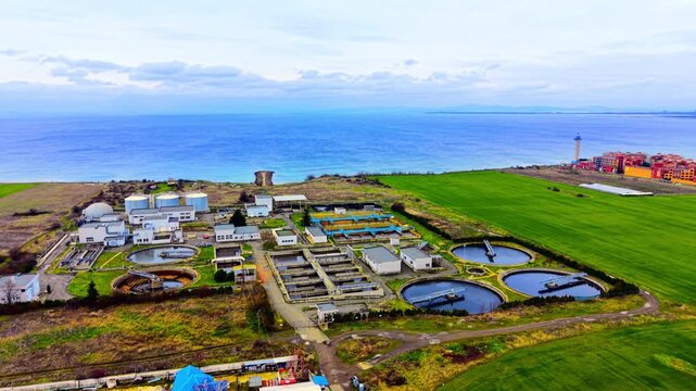 Aerial view of coastal wastewater treatment plant with Black Sea horizon, Bulgaria. Wide drone perspective showing clarifiers, aeration basins, digesters, green coastal fields and blue sea beyond.