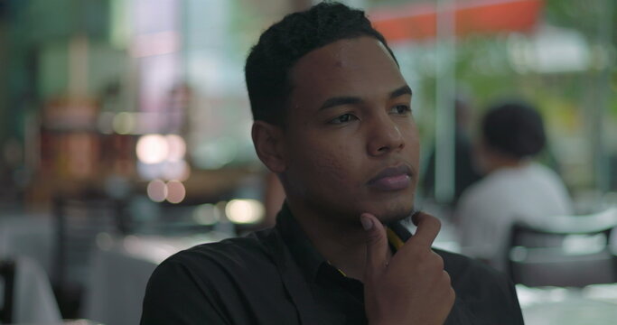 Color close-up of young restaurant staff member resting chin on hand in thoughtful pose, seated by window in quiet dining room