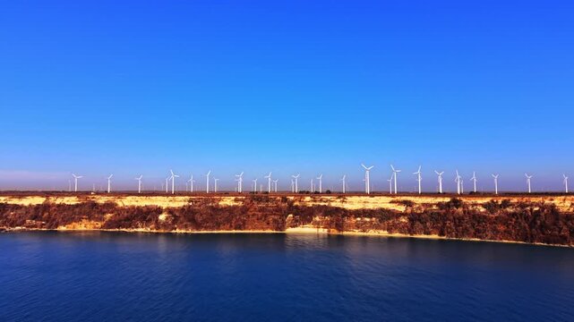 Wind turbines above limestone sea cliffs at the Black Sea, Bulgaria. Layered sandstone cliffs center, turbine row along plateau top, deep blue water below, clear sky above.