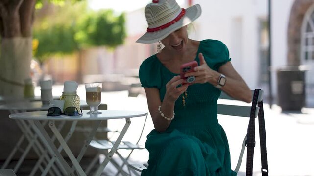 Happy senior digital nomad woman tourist looks at her phone and sips coffee at cafe near Loreto mission church while enjoying her time exploring Loreto in Baja California Sur, Mexico