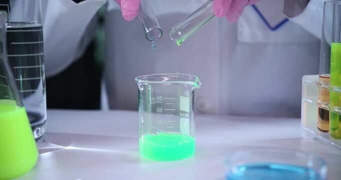Laboratory worker pours bright liquids from test tubes into beaker. Specialist slowly mixes colorful substances together in sterile laboratory
