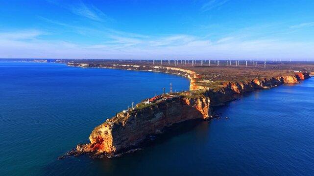 Cape Kaliakra peninsula and Black Sea, Bulgaria. Aerial view above shows red cliff promontory surrounded by deep blue and teal water, lighthouse buildings on grassy top, wind turbines in background.