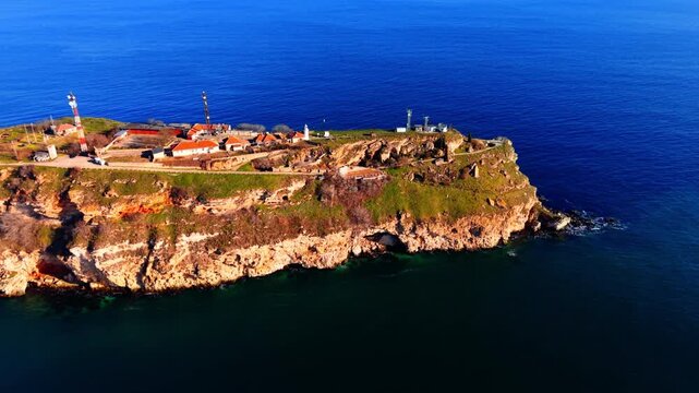 Drone view of lighthouse station on Cape Kaliakra cliff, Bulgaria. Red-roofed buildings, antenna towers, green scrub on orange cliff top, deep blue open sea fills frame.