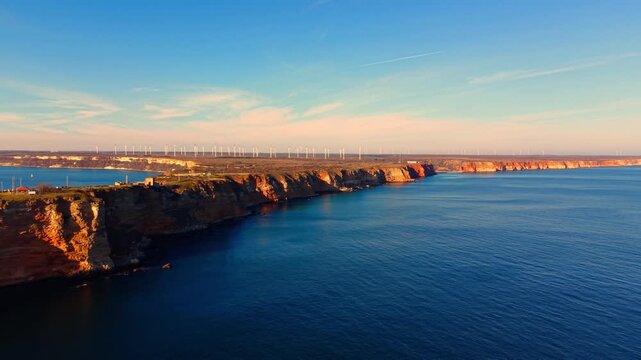Warm light on coastal cliffs and wind farm at dusk, Bulgaria. Aerial view with orange lit cliffs left, calm blue sea center, distant turbine row on plateau, soft pastel sky above.