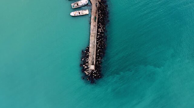 Concrete breakwater with moored boats. Directly aerial above perspective showing narrow jetty with tetrapod boulders, boats moored at tip, surrounded by turquoise water.