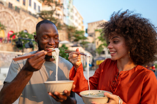 Young couple embracing the trend of Asian cuisine, happily eating noodles with chopsticks outdoors