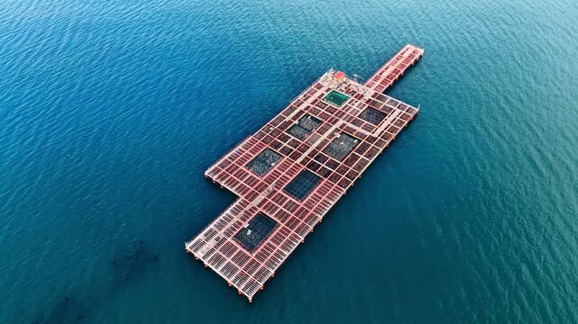 Aerial view of floating fish farm cages in deep blue sea, Bulgaria. Large red metal grid aquaculture platform with net pens on calm teal water.
