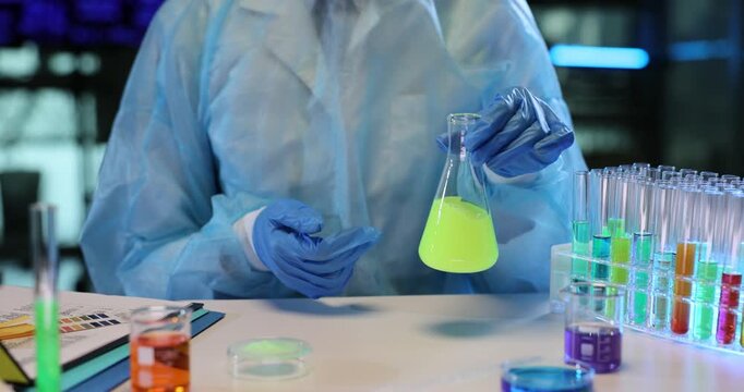 Laboratory assistant shakes bright radioactive liquid inside transparent beaker. Scientific worker in protective laboratory clothing mixes solution