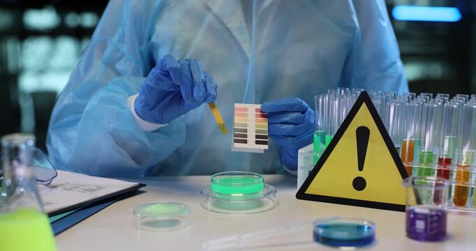Chemist drips strip of litmus paper into bright colorful liquid. Radioactive hazard warning sign stands clearly on laboratory table slow motion