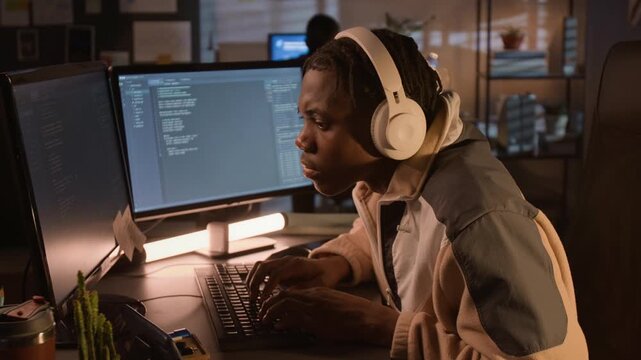 Young African American man wearing headphones typing code on computer in office at night. Footage represents software development, cyber security, and IT education marketing