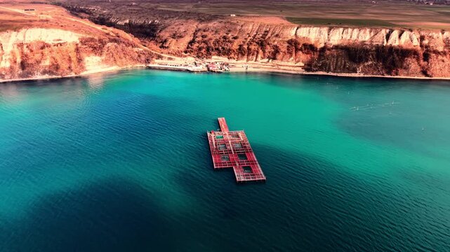 Fish farm platform in coastal bay with white chalk cliffs. Aerial view of red aquaculture structure on vivid turquoise water enclosed by eroded limestone headland and farmland above.
