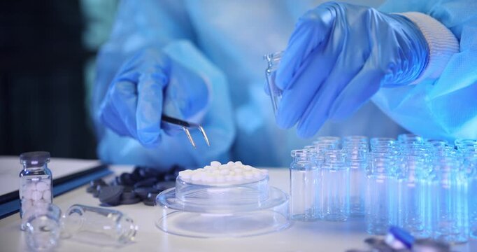 Lab worker uses metal tweezers to place small white tablets into tiny bottles. Scientific specialist prepares materials for medical analysis