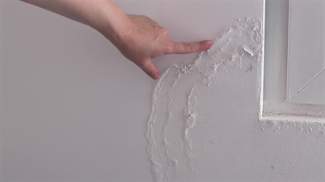 Water Damaged Wall Near Window. Person pointing at damaged plaster and peeling paint on an interior wall near a window. Moisture damage, home repair and renovation concept.