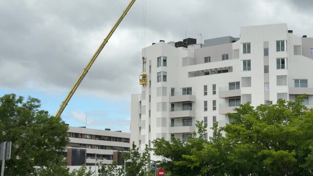 Construction crane operating next to a modern residential apartment building under a cloudy sky. Urban development and contemporary housing architecture in a city environment.
