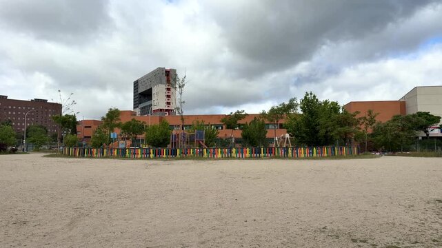 Madrid, Spain, June 9-2026: Wide view of an empty school playground with sandy surface, colorful fence, trees, and modern school buildings under a cloudy sky in an urban environment.