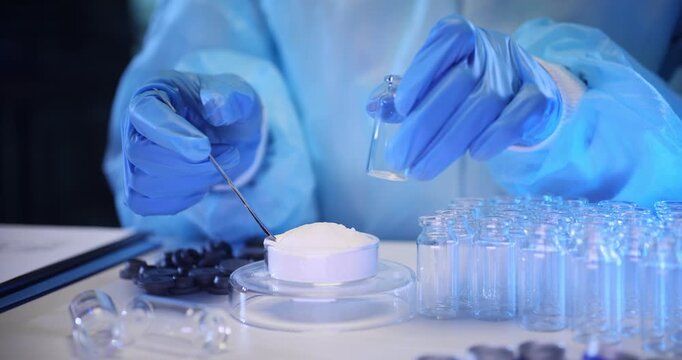 Lab technician transfers soft white powder into miniature glass containers. Worker uses laboratory spoon to place powder drug into tiny vials