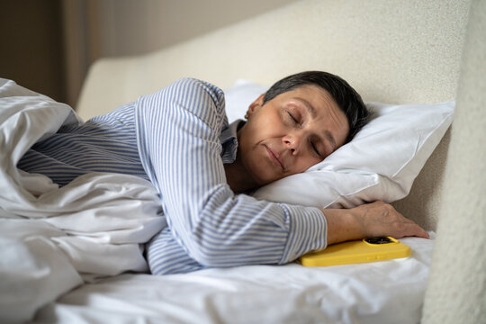 Calm senior woman comfortably sleeping in bed in at home with smartphone lying beside pillow, sleepy elderly female dreaming in morning procrastinating time in bed, gadget addiction and healthcare.