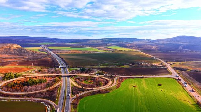 Highway interchange roundabout across green farmland valley. Aerial view of circular cloverleaf junction with trucks on road curving through colorful autumn fields toward distant hills.