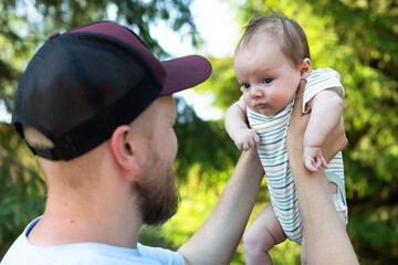 Naklejka premium Happy bearded father wearing a baseball cap holding his newborn baby high up in the air while smiling and looking at him in a sunny forest park