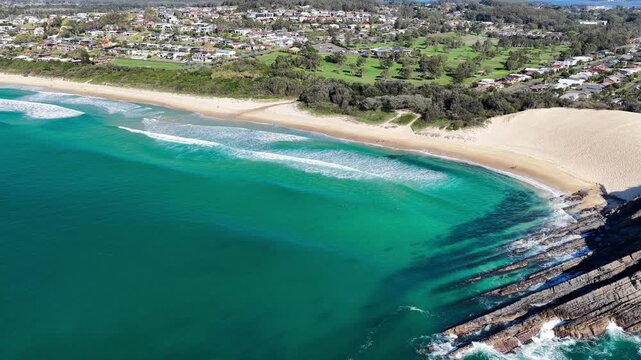 Aerial video flying over Bennetts Head at One Mile Beach near Forster, NSW, Australia, before descending towards the shoreline and following the rolling waves as they break onto the sand.