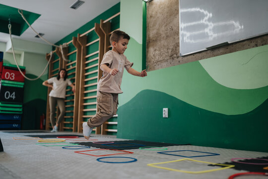 A young boy jumps through colorful hoops on the floor of an indoor gym during a physical education activity.