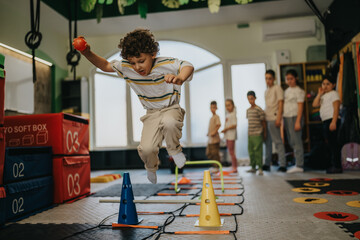A young boy jumps over cones while holding a ball during a kids fitness obstacle course. Other children watch in the background as he practices agility and balance in an indoor gym. © qunica.com