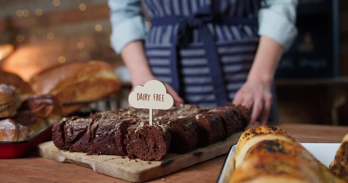 Female Sales Assistant In Bakery Putting Dairy Free Label Into Tray Of Brownies