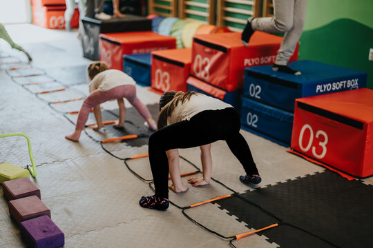 Young children doing agility ladder and balance exercises in a bright indoor gym. The group practices kids fitness drills using plyo boxes and coordination ladders.