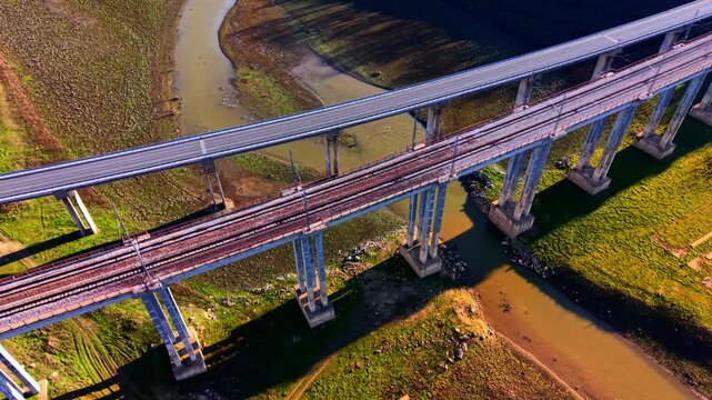 Aerial view of parallel road and railway bridges. Elevated perspective looking down at concrete viaduct pillars crossing a shallow river bed surrounded by green grass.