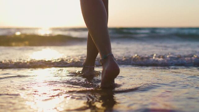 A rear view of a girl's legs and bare feet as she walks along the shoreline on wet sand during golden hour, with gentle waves, warm backlighting and tranquility in the background.