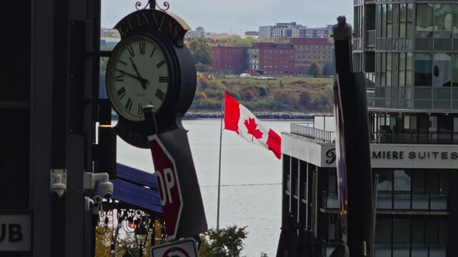 A flag of Canada flutters in Halifax, Nova Scotia against the backdrop of the waterfront. A clock tower shows the time as the day unfolds with overcast skies