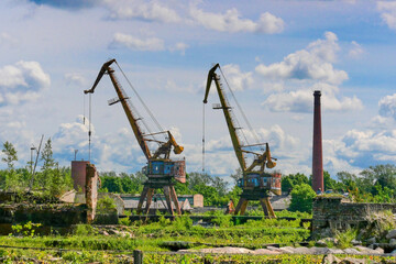 Fototapeta premium Abandoned rusty portal cranes and an old industrial brick chimney at a ruined shipyard under a cloudy summer sky.