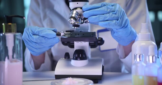 Doctor looks at cream specimen in microscope on lab desk. Dermatologist conducts research of innovative rejuvenation products in cosmetic laboratory