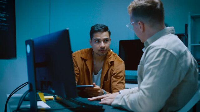 Two young male IT specialists discussing computer software programming code and web design development while working together in modern corporate office room with computers