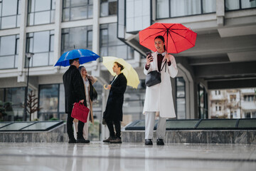 A businesswoman holding a red umbrella checks her smartphone in a rainy urban plaza. Three colleagues stand nearby under colorful umbrellas outside modern office buildings. © qunica.com