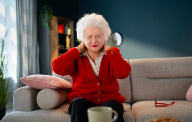 Obraz premium An elderly woman is seated on a sofa in a living room, holding her neck with both hands. She appears to be in discomfort. A cup and some cushions are nearby.