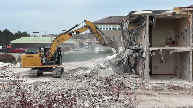 A construction machine is demolishing a building while school buses are parked nearby in Halifax, Nova Scotia during the daytime. Dust and debris fill the air