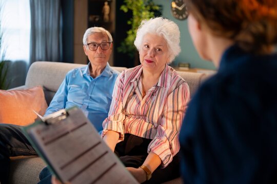 An older couple sits on a couch in a cozy living room. They are having a discussion with a person holding a clipboard. The room is bright and has plants and a clock on the wall.