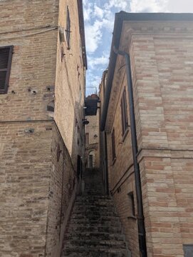 Una scalinata in pietra sale tra edifici storici in mattoni, guidando lo sguardo verso un piccolo arco illuminato dal cielo, in un tipico scorcio suggestivo dei borghi europei