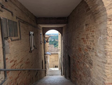 Un vicolo storico in mattoni scende verso un arco panoramico che si apre su tetti antichi e paesaggi verdi, creando un&rsquo;atmosfera autentica e suggestiva tipica dei borghi tradizionali italiani
