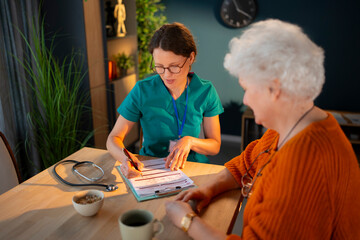 A nurse sits at a table discussing health matters with an elderly woman. They look at papers while...