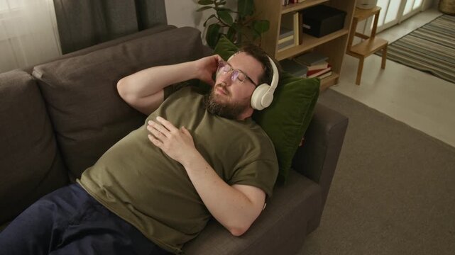 High angle shot of adult Caucasian man with diabetes lying on sofa and listening to music in headphones. Footage ideal for mental health apps, stress relief campaigns, and audio streaming services