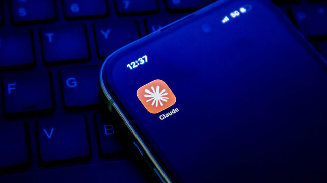 Closeup of a smartphone screen displaying the claude ai app icon on a dark background with a computer keyboard visible in the background