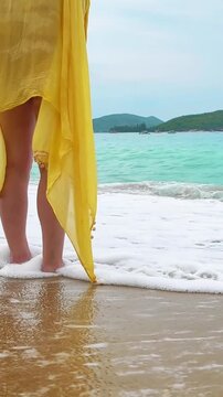 Female legs in yellow pareo standing at shoreline. Foamy ocean waves wash over bare foot. Horizon line in background. Wind blows pareo fabric. Tropical beach scene, relaxing seaside.