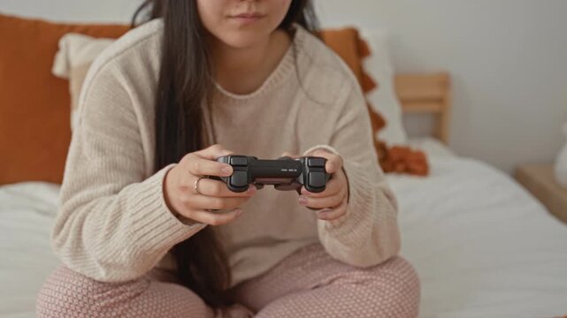 Woman holding game controller with thumbs on joysticks, sitting cross legged on bed in bedroom; concentration leisure.