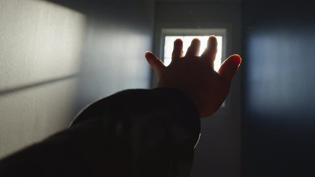 A hand extends toward a small opening that lets in light in a dark room. The warm light shines through as the person tries to reach it