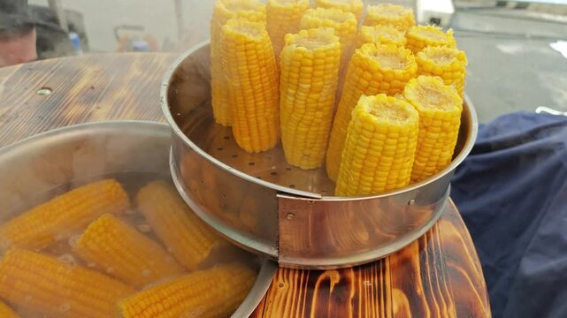 Person in black gloves lifts steaming corn cobs from a metal steamer, showcasing the vibrant yellow kernels and wooden table surface in a food preparation setting