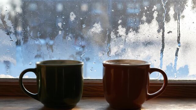 Two steaming mugs of hot beverage on a windowsill with frosty ice patterns on the glass