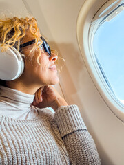 Naklejka premium Woman with headphones and sunglasses looking out airplane window, relaxing during a flight in a cozy sweater and turtleneck, enjoying travel, calm, and in-flight comfort