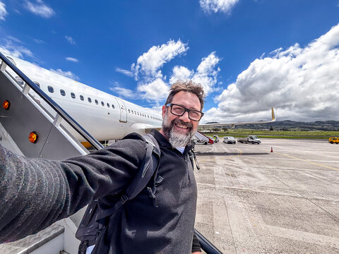 Happy man taking a selfie on airport tarmac while boarding a plane, smiling with backpack under blue sky and clouds, travel, adventure and business trip concept